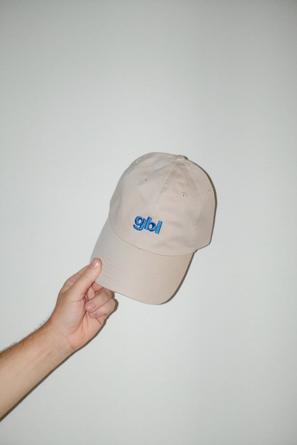 Beige cap with 'gbl' logo held by a hand against a plain background