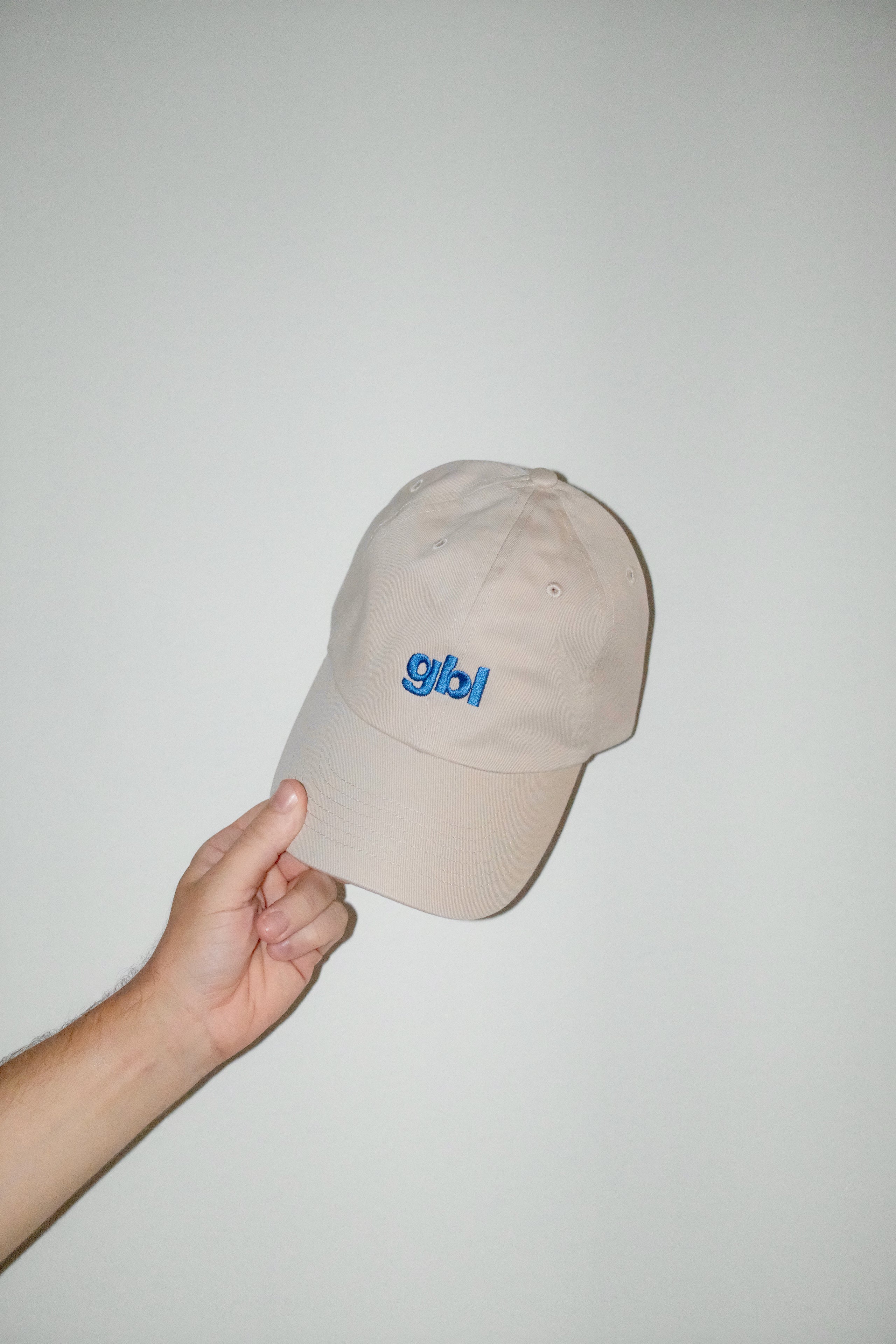 Beige cap with 'gbl' logo held by a hand against a plain background